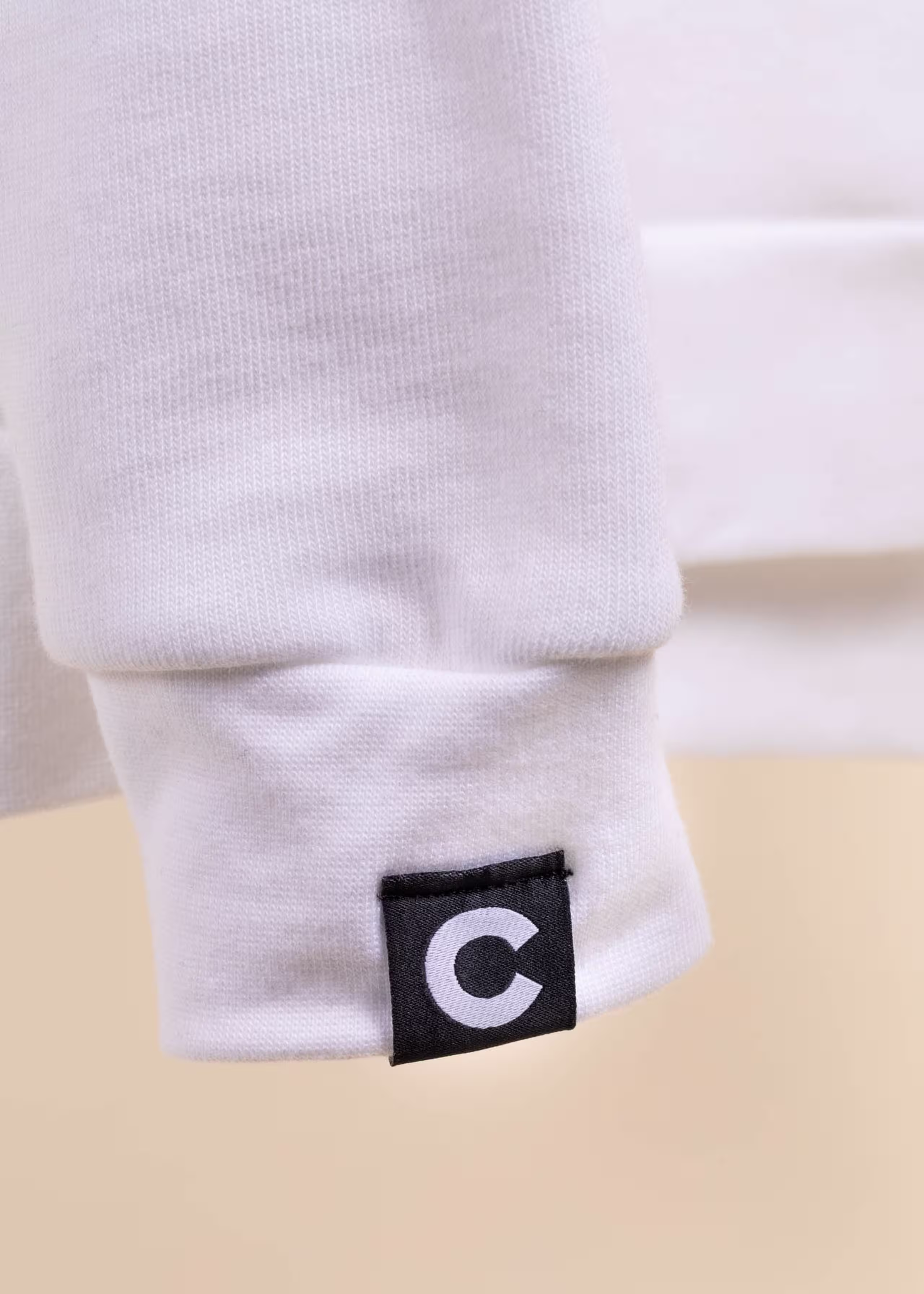 A black hem tag with a white "c" on it is attached to the sleeve of a white sweatshirt